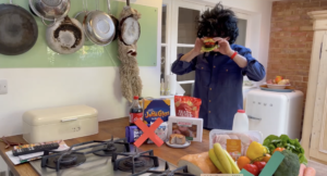 Phil, in disguises wearing a big black bushy wig, is caught biting into a big burger. In front of him is two groups of food. In front of one group, consisting of coca-cola, Jaffa Cakes, crisps etc is a big red cross, in front of the other group of fruit and veg, is a green tick