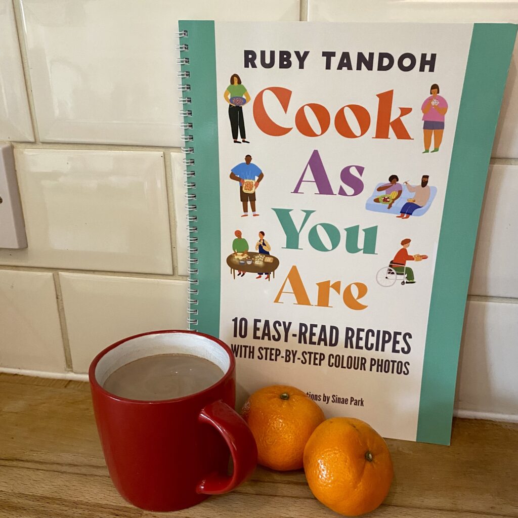 The easy read version of cook as you are, propped up, with a mug of clementine hot chocolate and some clementines in front of it
