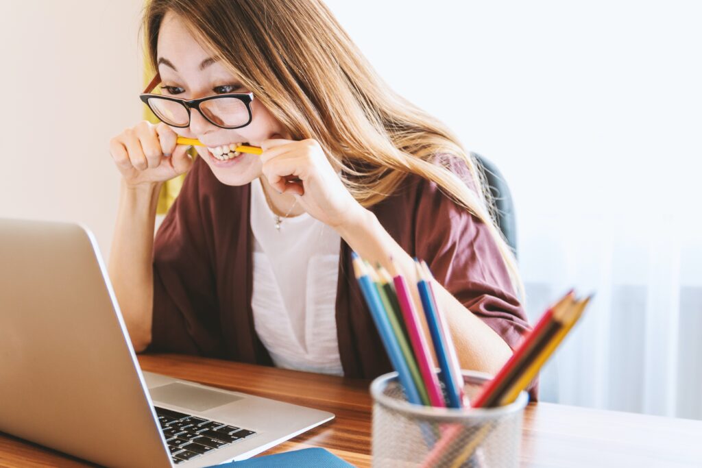 A woman sits at a desk in front of an open laptop, a pencil pensively between her teeth