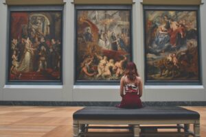 A woman, sitting on a museum bench, with her bck to the camera, looking at large, old paintings