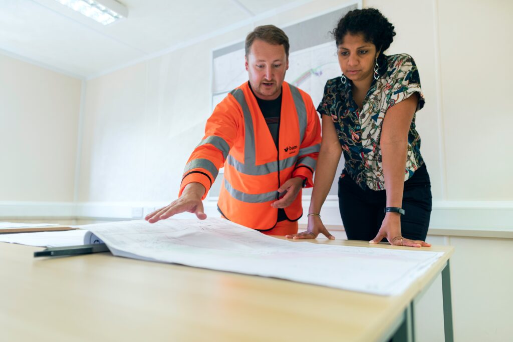 Two people, one in a hi-vis jacket, stand looking at building plans