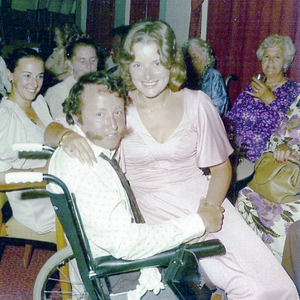 A vintage photo. Jessica sits on Wes's (who is a wheelchair) lap. They are at a party or celebration.