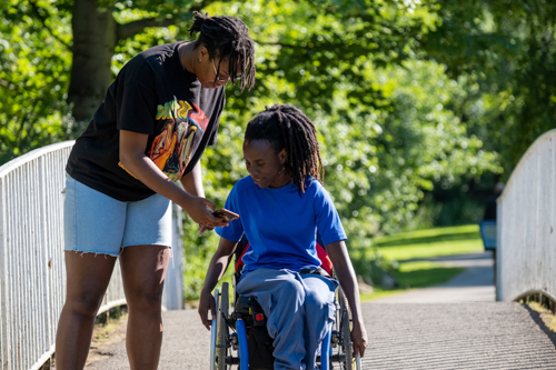 Teenage girl (16-17) in wheelchair with friend walking in park