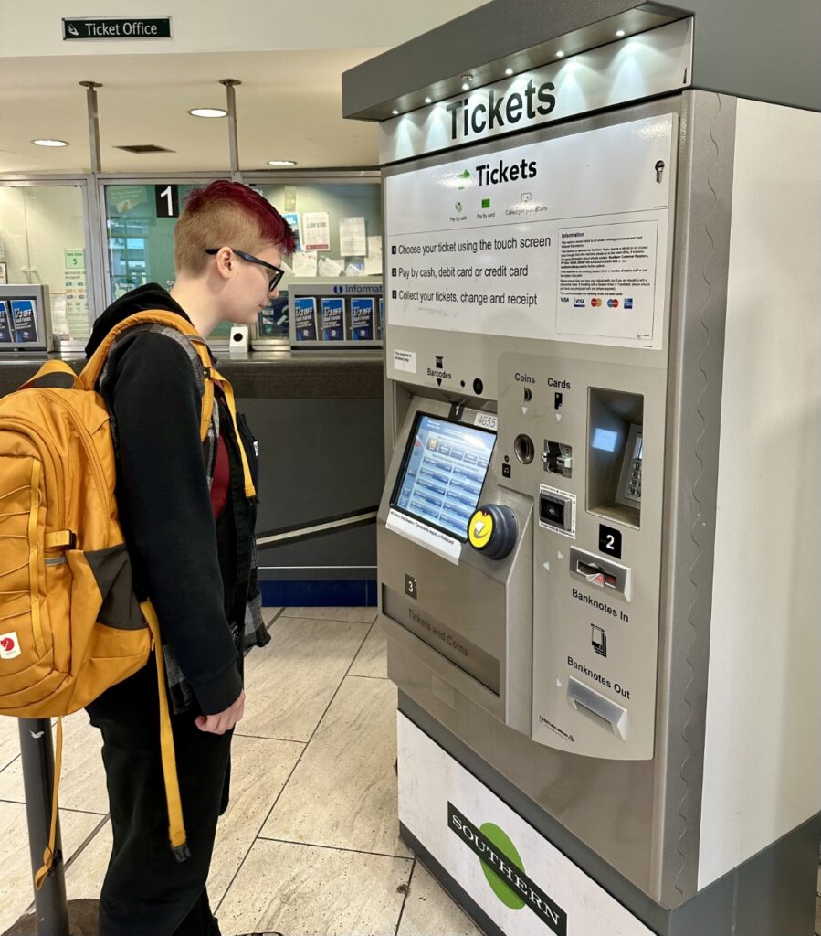 A young person stands looking at a train station ticket machine