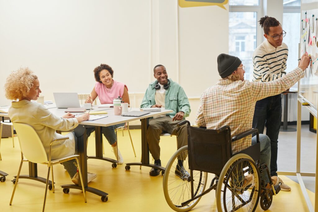 People around a table at a meeting, a person in a wheelchair is writing something on a whiteboard