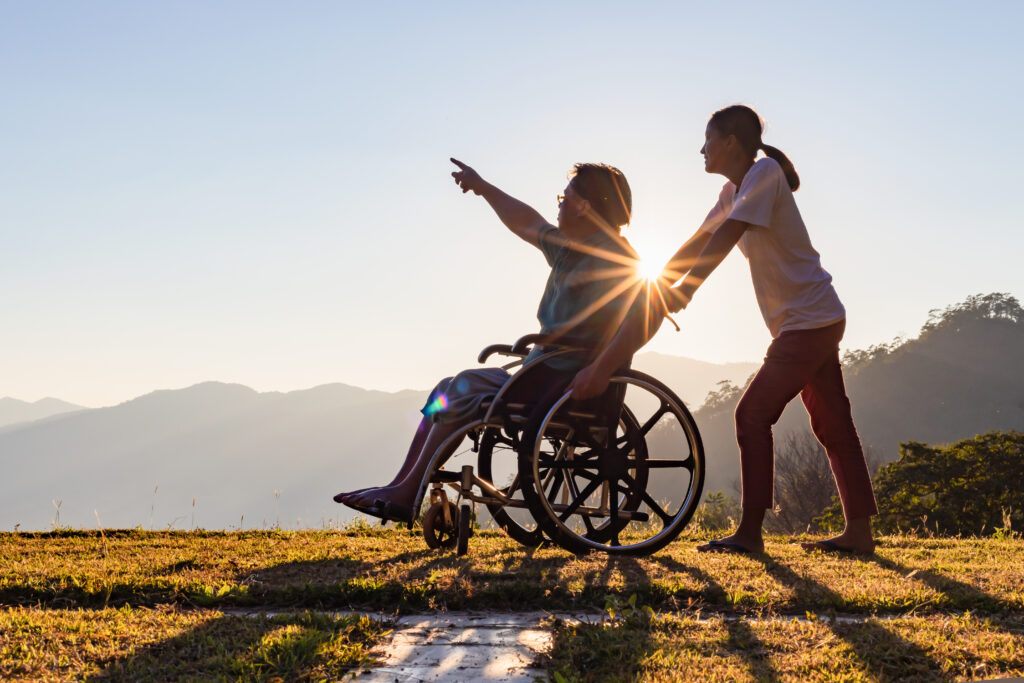 A woman pushes a man in a wheelchair outside in what looks like an area of beauty, the sun is low in the sky, there's mountains in the distance. The man is pointing at something in the distance.