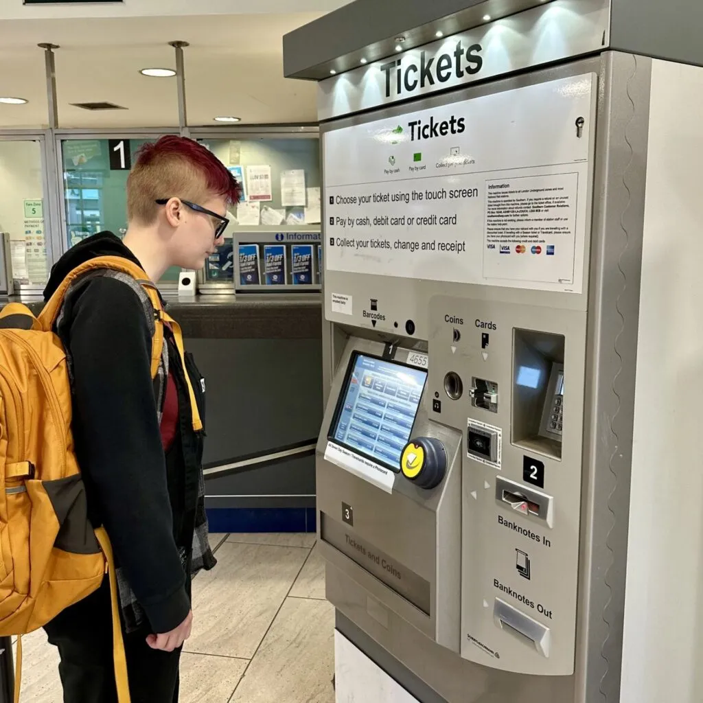 A young person stands looking at a train station ticket machine