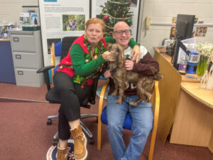 Aggie and Jason with a wiggly dog, sitting in front a Christmas tree in the KCIL office, they are both wearing Christmas jumpers, Aggie's jumper is covered in actual Christmas decorations