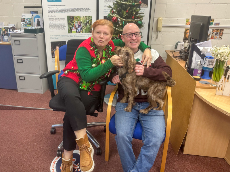 Aggie and Jason with a wiggly dog, sitting in front a Christmas tree in the KCIL office, they are both wearing Christmas jumpers, Aggie's jumper is covered in actual Christmas decorations