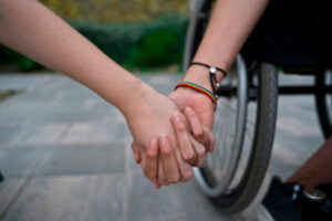 Soft focus of crop anonymous female holding hands with girlfriend wearing LGBT wristband using wheelchair during summer day spending time together